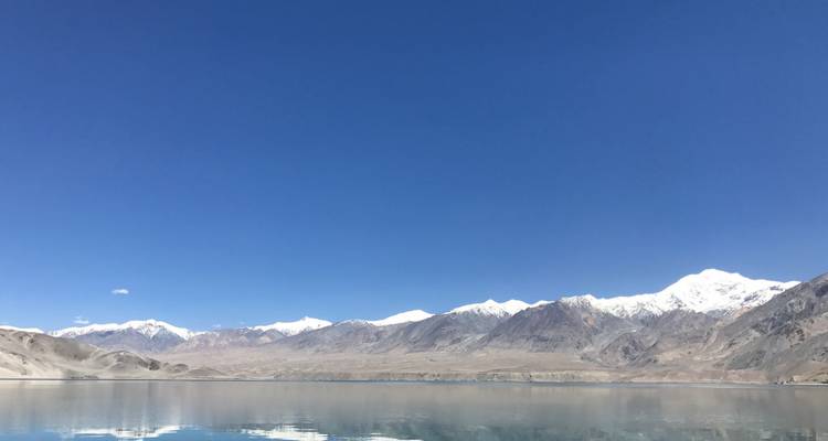 Wide panorama of barren mountains with snowy summits mirrored in a calm lake.