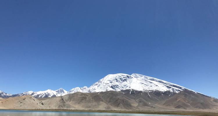 Prominent snow-covered Muztagh Ata towering over arid foothills and turquoise lake.