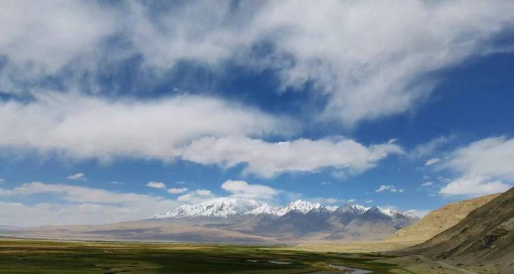 Expansive green valley with winding river framed by snow-capped mountain range and dramatic clouds.