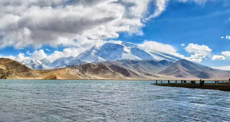 Turquoise lake bordered by barren hills and lofty white peaks under dramatic clouds.