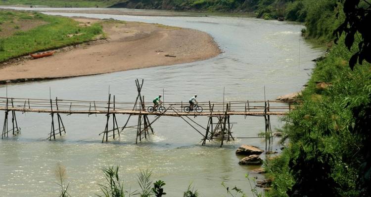 Deux cyclistes traversant un pont en bambou rustique enjambant une rivière sinueuse au milieu d'un paysage verdoyant.