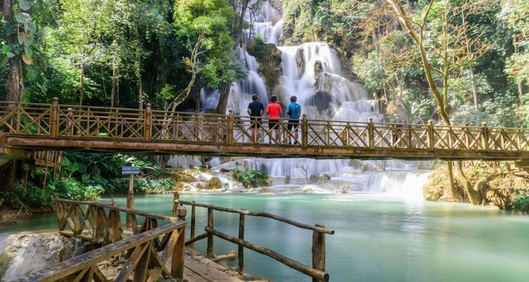 Trois cyclistes se tenant debout sur un pont en bois admirant une cascade à plusieurs étages dans une forêt dense.