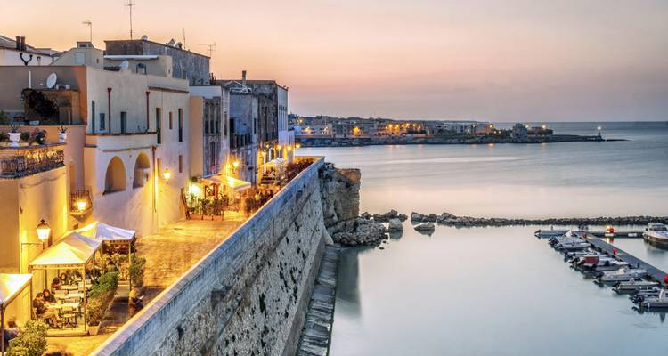 Ancienne ville côtière avec remparts en bord de mer, lumières de café et eaux calmes du port au coucher de soleil doux