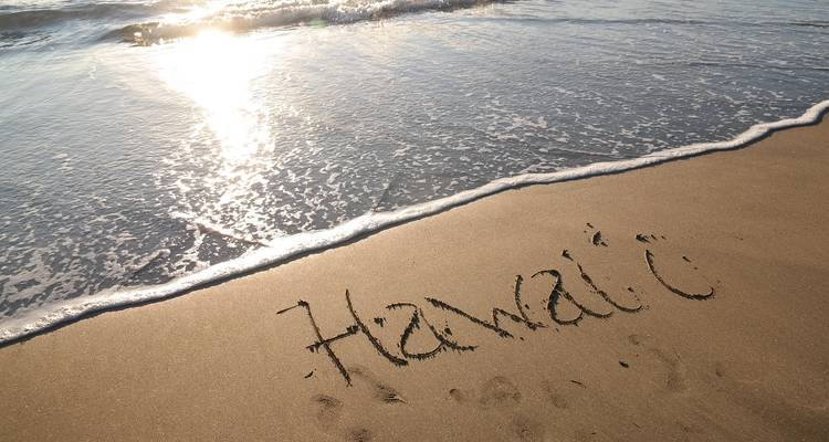 Sandy beach with gentle wave and sunlight, the word “Hawaii” written in the wet sand.