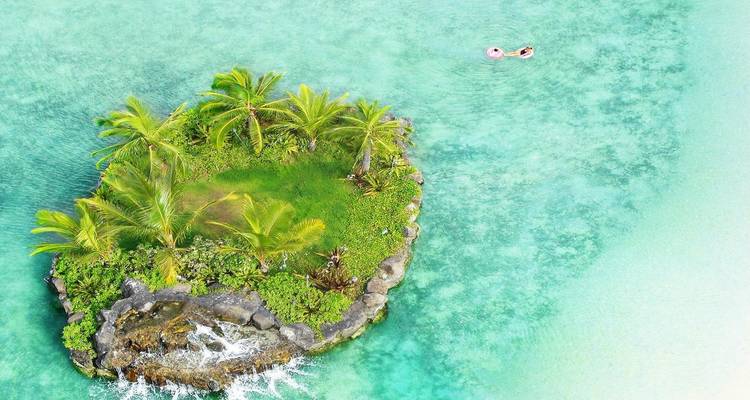 Tiny palm-covered islet surrounded by crystalline turquoise water with two swimmers on floaties nearby.
