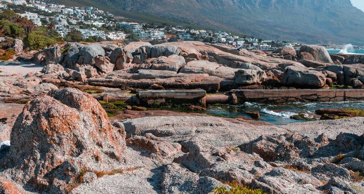 Des rochers de granit accidentés le long du rivage avec des maisons côtières lointaines et un arrière-plan montagneux sous une lumière diurne éclatante.