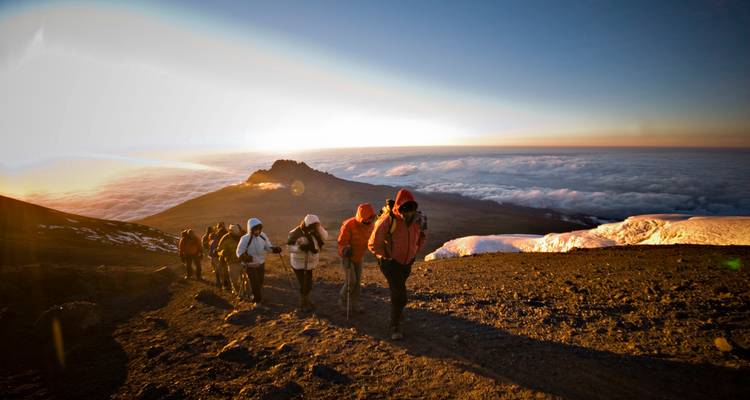 Group of climbers in colorful jackets trekking up a volcanic slope toward a glowing sunrise above cloud layers