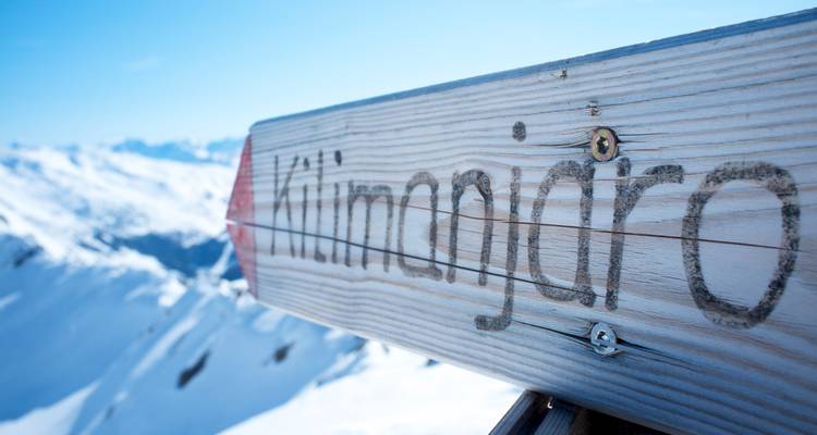 Weathered wooden sign reading “Kilimanjaro” points toward snowy alpine peaks under clear sky