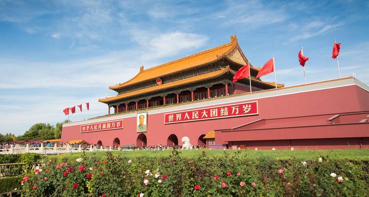Tiananmen-Tor mit roten Mauern, Flaggen und Porträt, umrahmt von blühenden Rosen unter blauem Himmel.