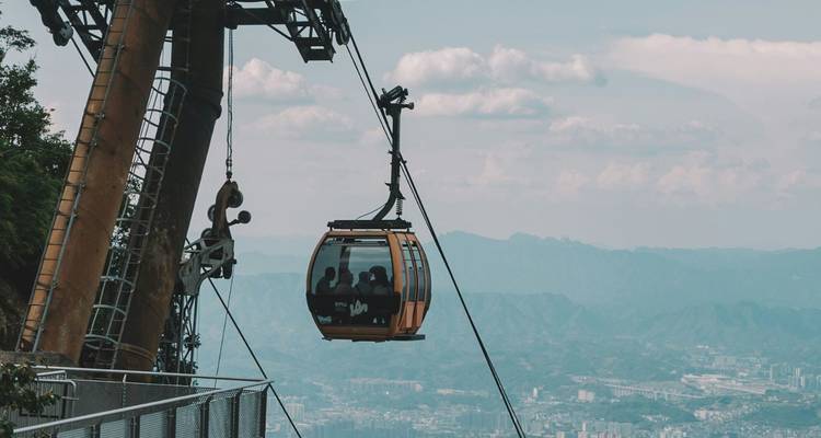 Geschlossene Seilbahn, die über eine dunstige Berglandschaft aufsteigt, Fahrgäste sind durch Glas sichtbar.