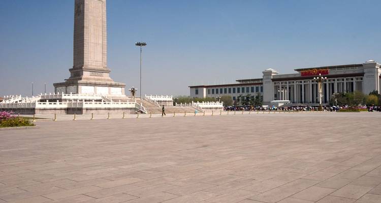 La inmensa Plaza de Tiananmen pavimentada con el Monumento a los Héroes del Pueblo y el Museo Nacional bajo un cielo azul despejado, con una multitud dispersa a la distancia.