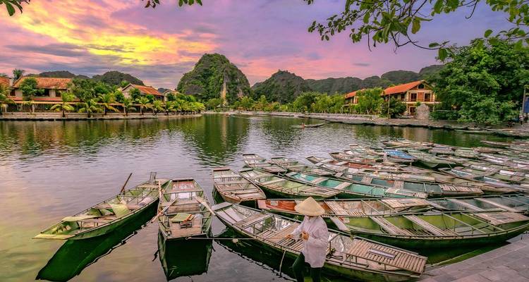 Des barques alignées le long d'une rivière tranquille au coucher de soleil coloré tandis qu'une femme portant un chapeau conique attend près de l'eau.