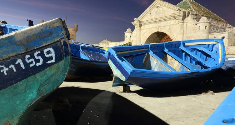 Blue wooden fishing boats rest on the harbor sand near Essaouira’s fortified gate at dusk.