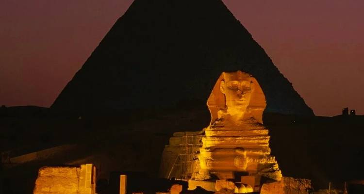 De Sfinx verlicht 's nachts met de donkere silhouet van een piramide op de achtergrond.