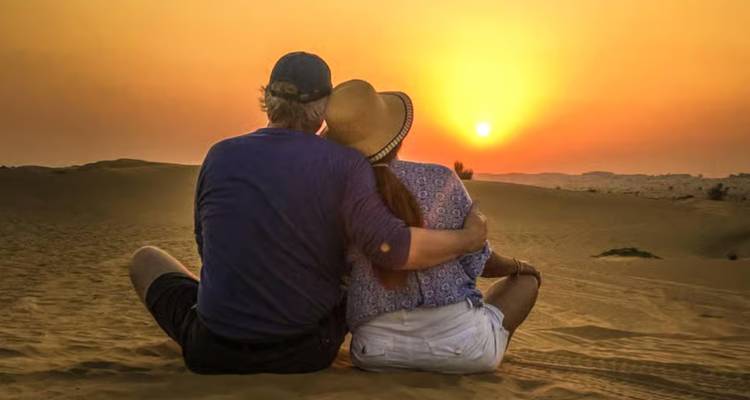 Ein Paar sitzt Arm in Arm auf dem Wüstensand und beobachtet einen goldenen Sonnenuntergang in der Sahara.