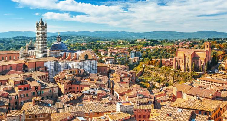 Colorful skyline of Siena with its striped cathedral tower and medieval brick buildings under blue skies.