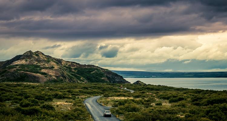 La route côtière sinueuse mène vers les eaux lointaines sous des nuages dramatiques et des collines escarpées.