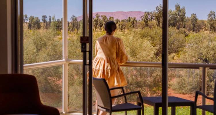 Un client s'appuie sur la balustrade d'un balcon et regarde vers Uluru à travers le paysage désertique.