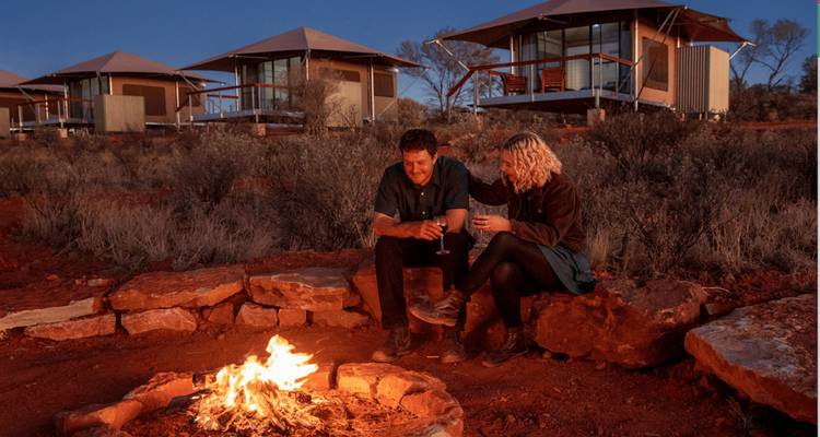 Un couple déguste du vin près d'un feu de camp devant des cabanes de luxe sous un ciel crépusculaire.