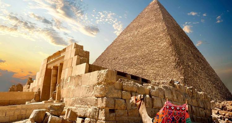 Iconic Great Pyramid of Giza rises under a dramatic sunset sky with ruins and a colourful camel in the foreground.