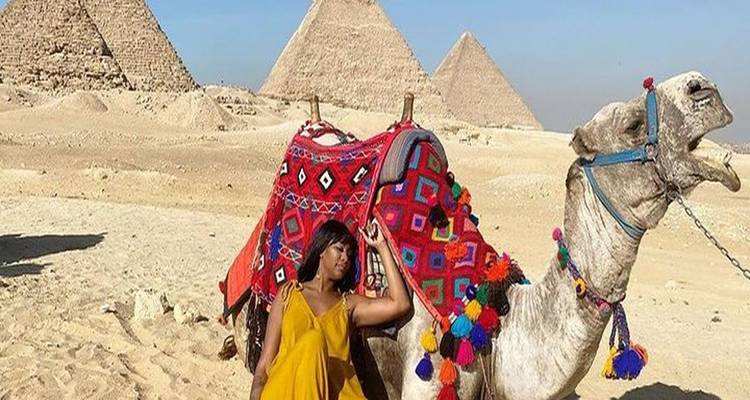 Woman in a yellow dress poses beside a decorated camel with the pyramids rising out of the desert behind.