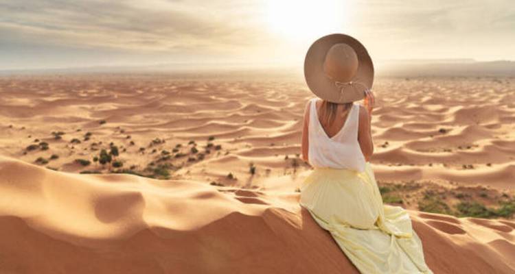 Woman in a flowing yellow skirt sits on a high dune gazing over endless Sahara sands at sunrise.
