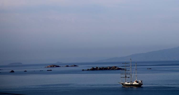 Tall-masted sailing ship gliding across calm blue sea with hazy islands beyond