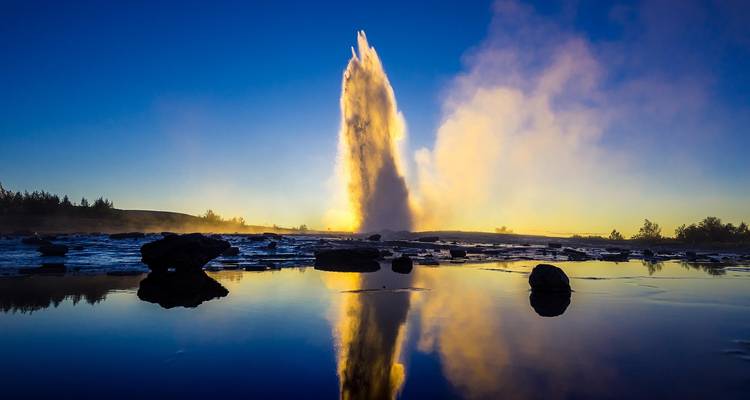 Un poderoso géiser erupciona en el cielo azul del atardecer, reflejándose perfectamente en un tranquilo estanque de agua