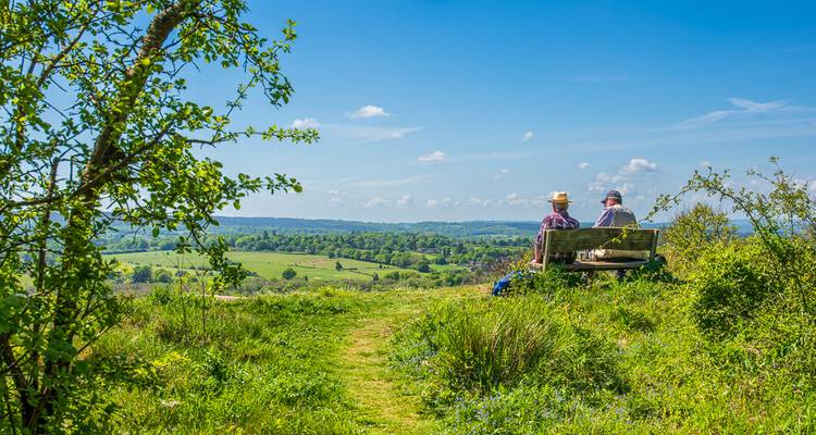 Zwei Personen entspannen sich auf einer Bank mit Blick auf die sanften grünen Hügel der Cotswolds-Landschaft