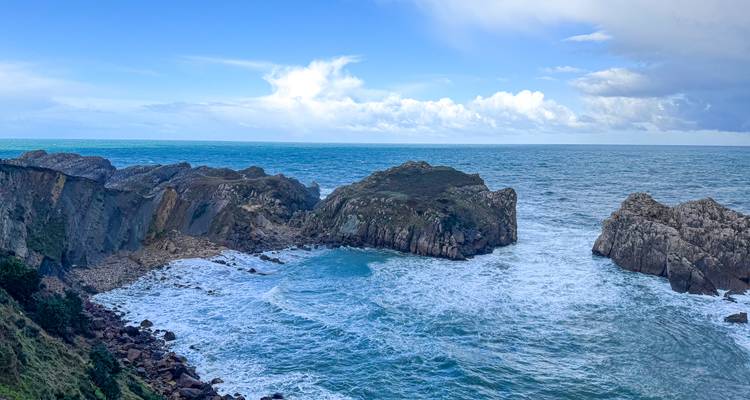 Rotsen aan de kust met golven die tegen een klein turquoise baaitje klappen onder een helder uitziende hemel.