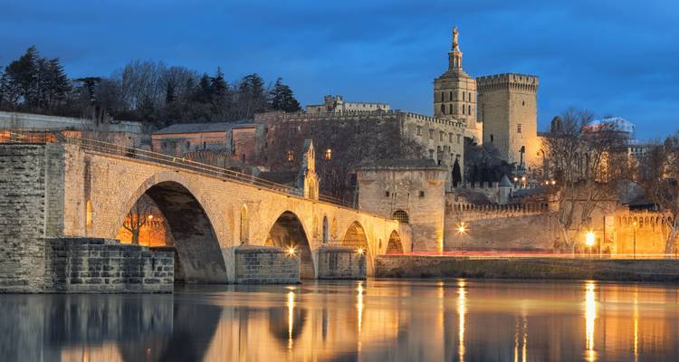 Illuminated medieval bridge and palace reflect on the Rhône River at dusk in Avignon.