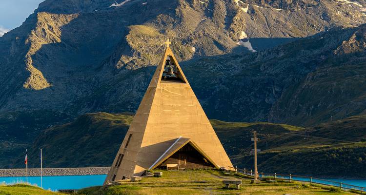 Triangular alpine chapel stands before dramatic mountains and a blue reservoir.