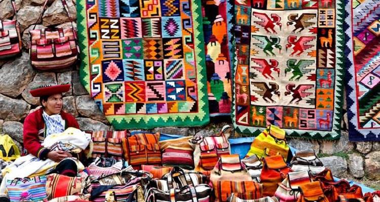 Woman in traditional attire sells vibrantly patterned textiles and bags at an Andean market stall.