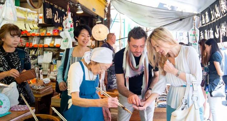 Travelers sample local snacks from an elderly vendor at a bustling traditional market stall.