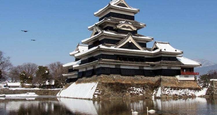 Snow-covered black-and-white castle reflected in a still moat with distant mountains.