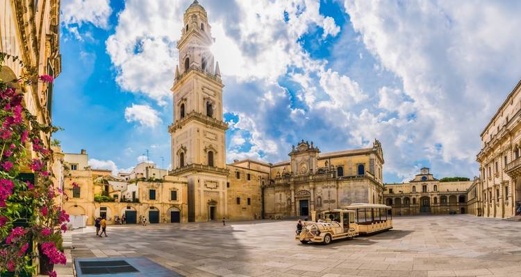 Vue panoramique de la place de la cathédrale baroque de Lecce avec un petit train touristique sous un ciel dramatique.