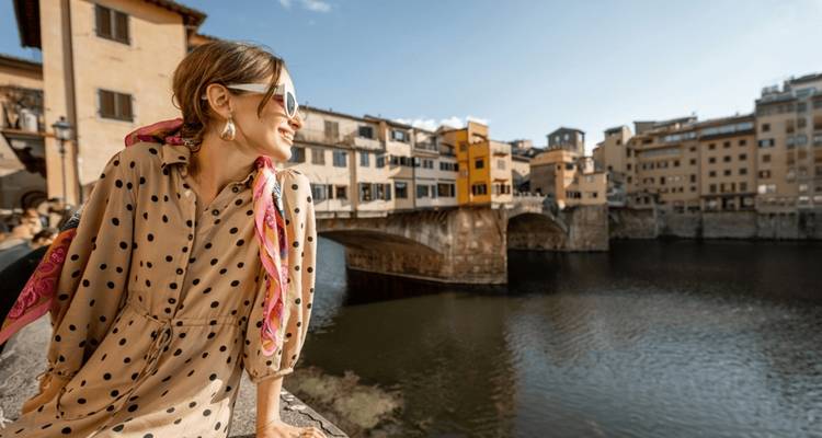 Stylish woman smiles while leaning on a riverside wall overlooking Florence’s Ponte Vecchio.