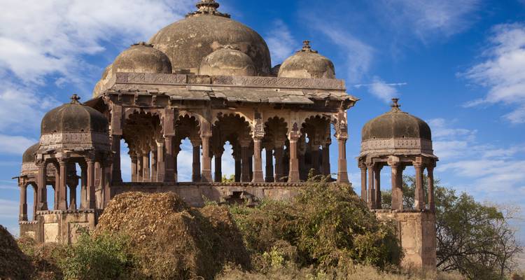 Cénotaphe en pierre patinée aux dômes et arches s'élevant au-dessus de la végétation broussailleuse contre un ciel bleu du Rajasthan.