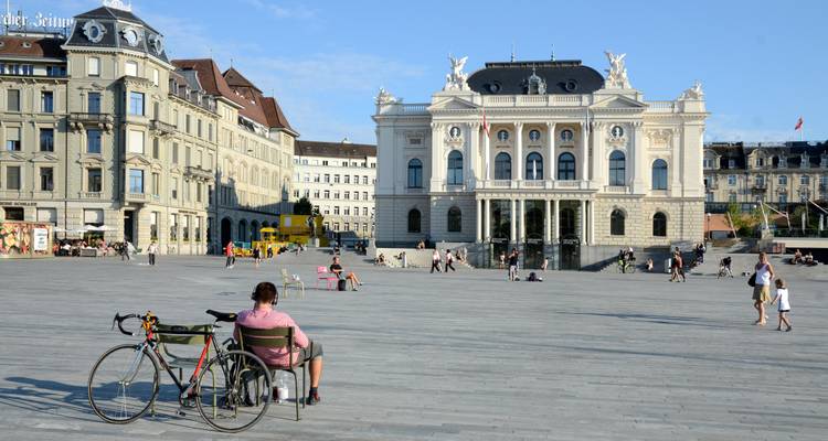 Plaza abierta frente a la Ópera de Zurich con ciclistas y peatones disfrutando de un día soleado.