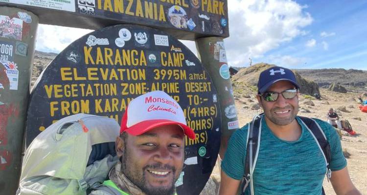 Two hikers pose beside a signboard at Karanga Camp showing elevation details on Mount Kilimanjaro.