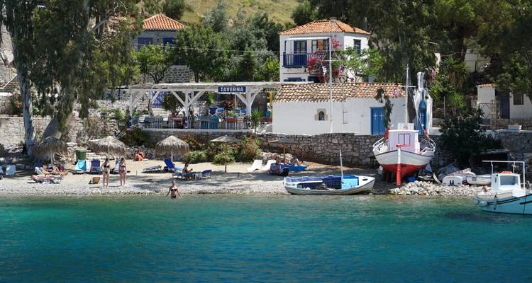 Small pebble beach with taverna and fishing boats on the clear turquoise water of Hydra.