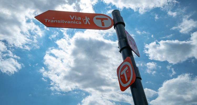Aufnahme von unten auf ein orangefarbenes Richtungsschild der Via Transilvanica, das über einen Himmel mit flauschigen weißen Wolken zeigt.