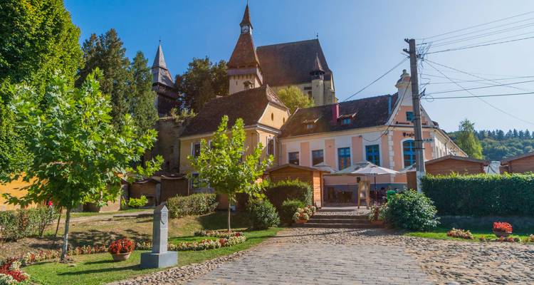 Gepflasterte Gasse mit Bäumen gesäumt und farbenfrohen Häusern, die zu einer befestigten Dorfkirche führt, mit Café-Gästen, die sich draußen entspannen.