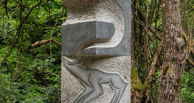 Steinpfeiler, der kunstvoll mit einem Tiermotiv geschnitzt ist und zwischen dichten grünen Waldbäumen steht.