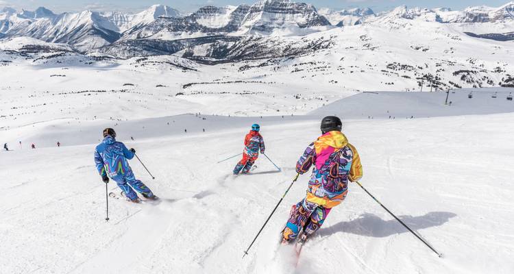 Drie kleurrijk geklede skiërs dalen af langs een brede besneeuwde bergflank