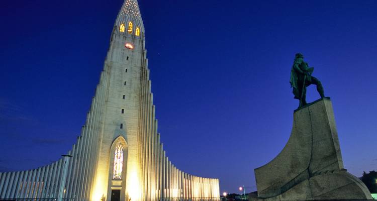Vue nocturne de l'église Hallgrimskirkja illuminée avec un ciel bleu profond en arrière-plan et une statue au premier plan.