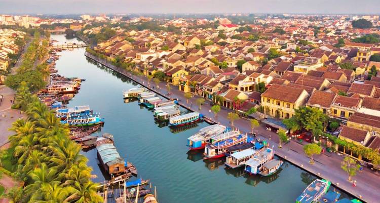 Una vista aérea de Hoi An muestra tejadillos alineados junto a un tranquilo río lleno de botes de turismo rodeados de palmeras.