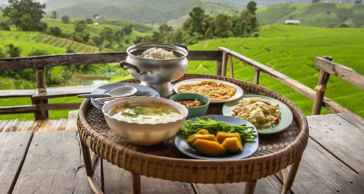 Una mesa de madera rústica que da a unas terrazas de arroz de un verde vibrante está puesta con arroz humeante, sopa y platos locales frescos.