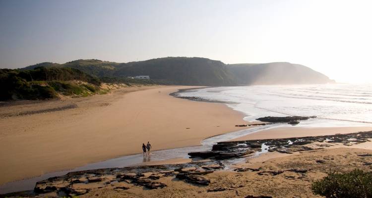 Vaste plage déserte avec deux promeneurs au loin, côte rocheuse et collines brumeuses en arrière-plan