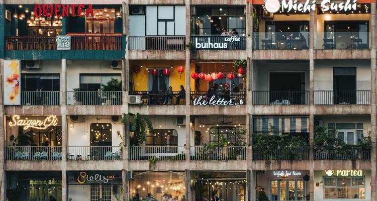 Ein mehrstöckiges Apartmentgebäude in Ho-Chi-Minh-Stadt, gefüllt mit trendigen Cafés und Boutique-Beschilderungen, wobei jeder Balkon mit Lichtern und Pflanzen dekoriert ist.